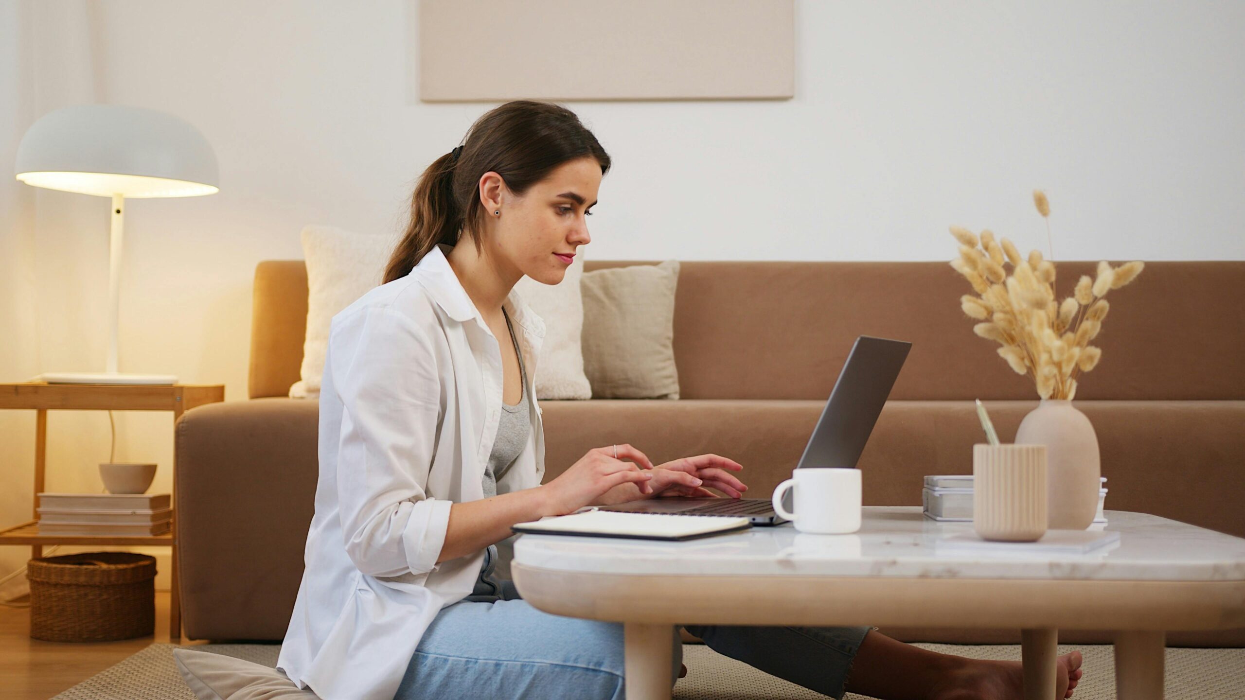 Side view of positive young lady with ponytail in casual outfit browsing contemporary netbook on coffee table while sitting on floor in cozy living room